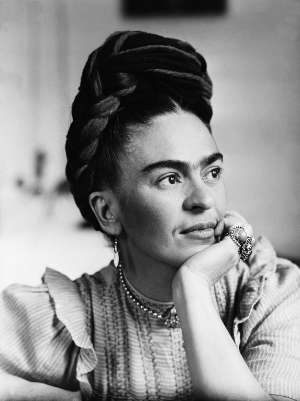 circa 1945:  Mexican painter Frida Kahlo (1907 - 1954) sits with her arms folded, looking down, in front of one of her paintings and a wooden bird cage. She wears flowers in her hair and a wooden necklace.  (Photo by Hulton Archive/Getty Images)